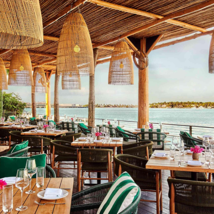 Patio al aire libre con luces colgantes de mimbre, techo de mimbre, pequeñas mesas de madera y sillas de estilo exterior con vistas a un mar azul.