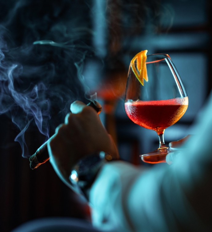 Hand holding a lit cigar and a cocktail with orange peel garnish at Smoke & Mirrors Doha