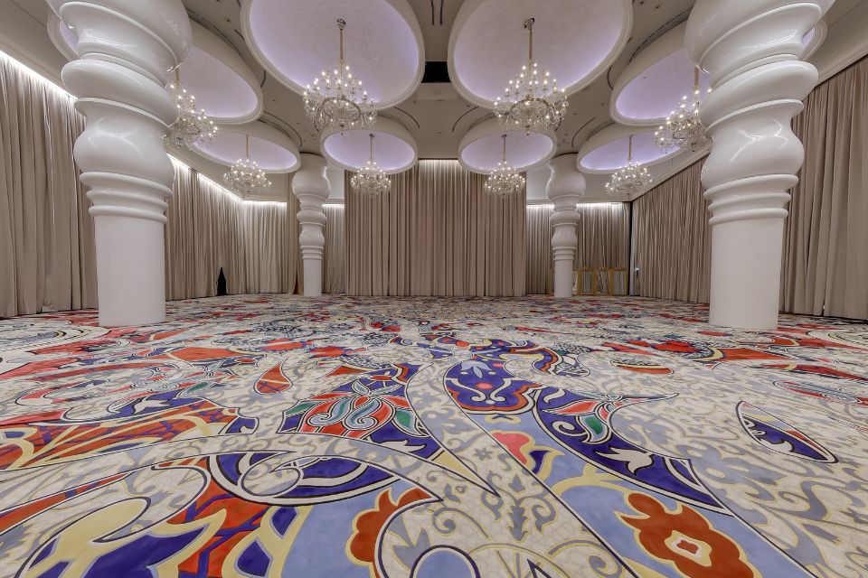 Large ballroom with white columns, patterned carpeted floors, grey curtains, and multiple chandeliers