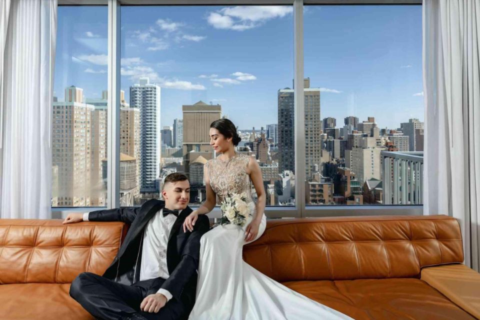 Bride and groom sit on a light brown leather sofa in front of a large window looking out to the New York City skyline