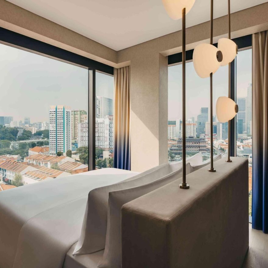 Large bed with cream headboard and modern lights above looks out to windows of cityscape