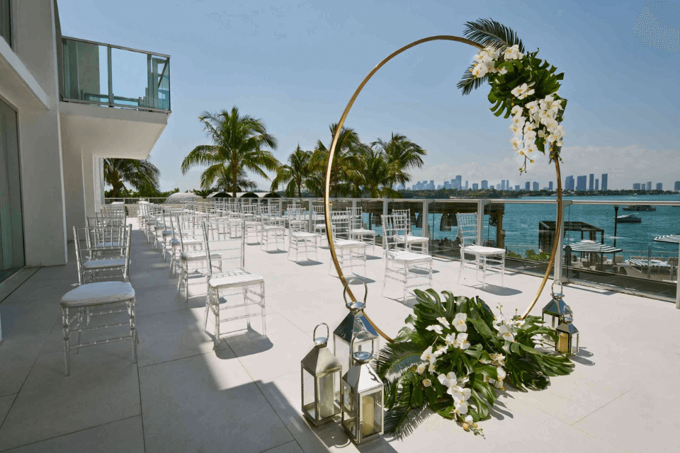 Outdoor wedding set up with rows of chairs, flower altar, and beach in the background with palm trees