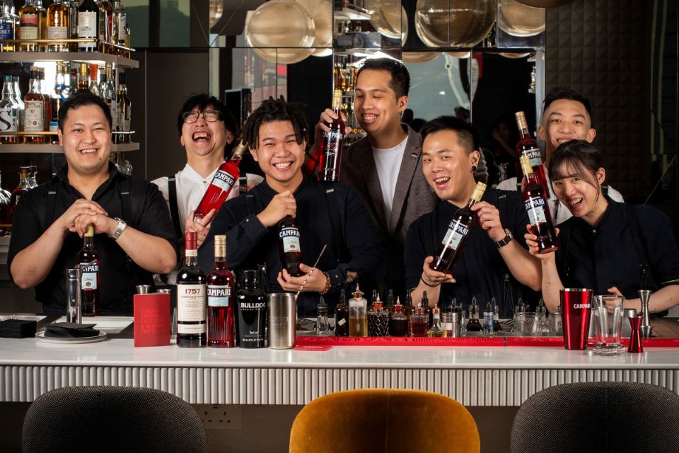 Multiple bartenders posing behind a bar with bottles of campari in their hands