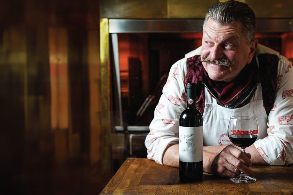 Man in chef's coat with glass of red wine and bottle of red wine on table in front of him