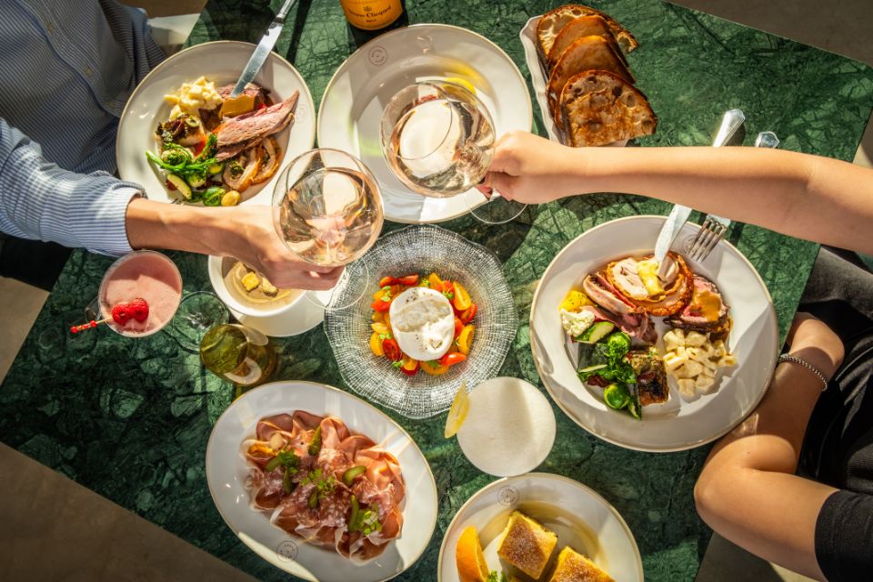 Multiple brunch dishes on a green tabletop with hands digging forks into plates of food