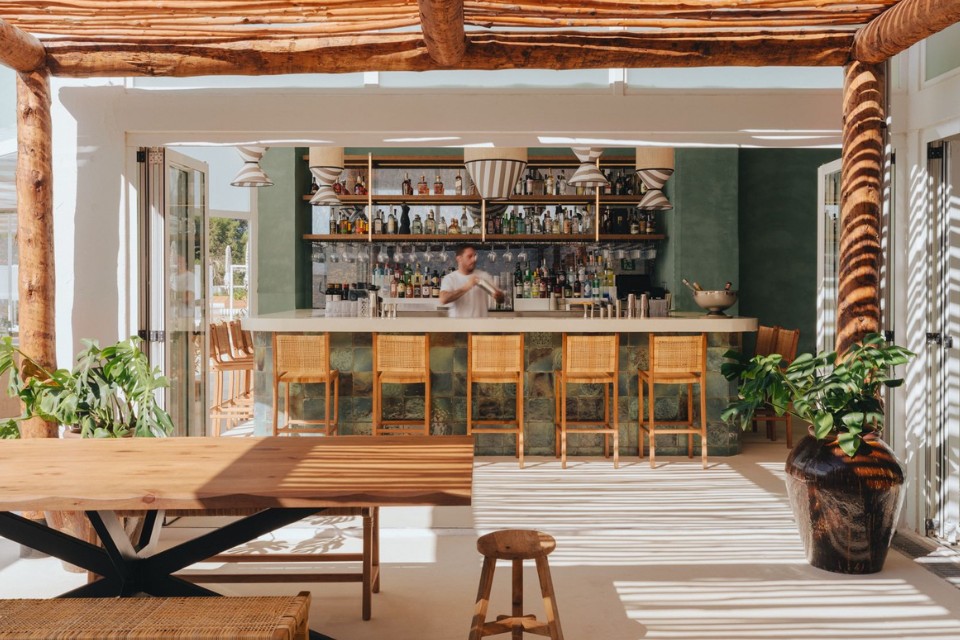 Boho-chic outdoor bar with light brown chairs and man behind bar shaking a cocktail