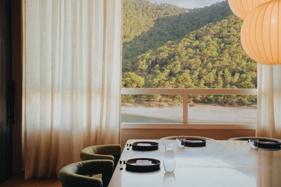 Modern dining table with two chairs, mulitple place settings, and paper chandeliers in the corner with a window overlooking a beach