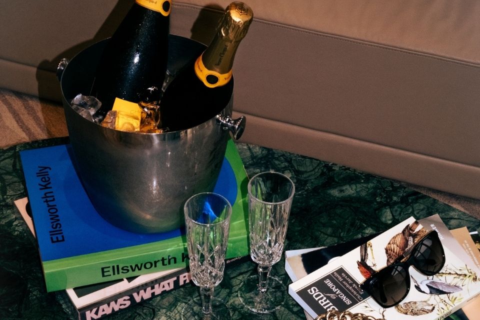 Two champagne bottles in a bucket on top of a stack of books with champagne flutes on a table and magazines and one pair of sunglasses