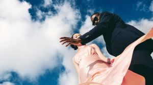 Man in dark suit and woman in pink dress pose in front of a blue cloudy sky