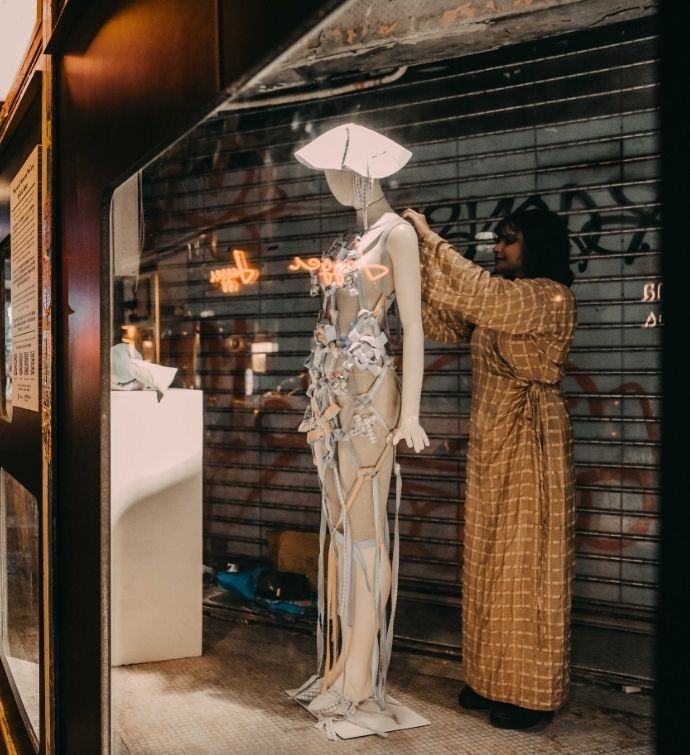 Woman in long beige dress stands behind a mannequin dressing it in a window front