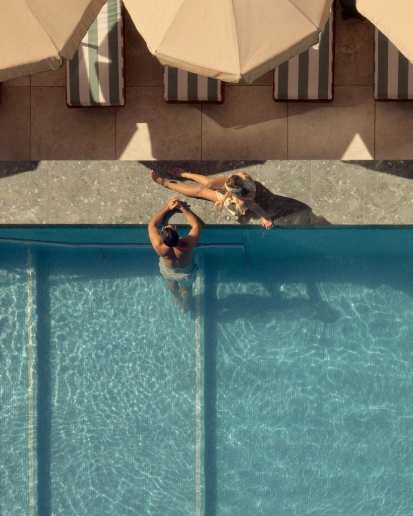 Blue pool with man in it and woman lounging beside it