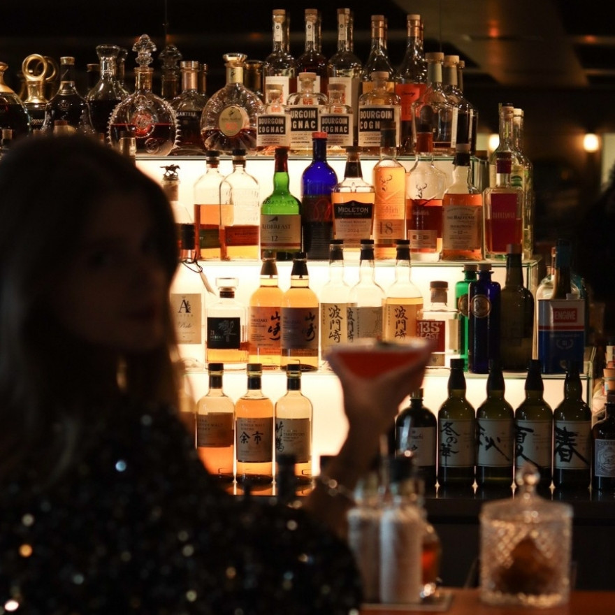 Lit up bar with shelves of liquor and shadow of woman holding a cocktail glass