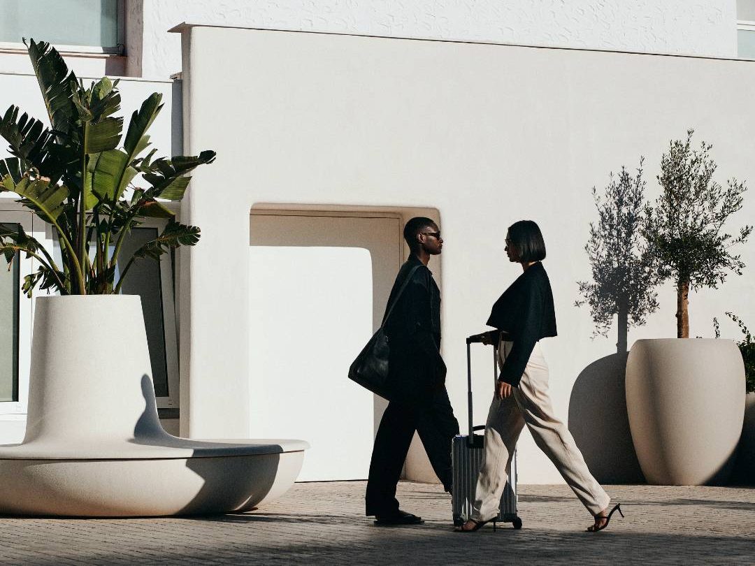 Man in black with bag and woman in black with white pants rolls a suitcase in front of a white building with palm trees