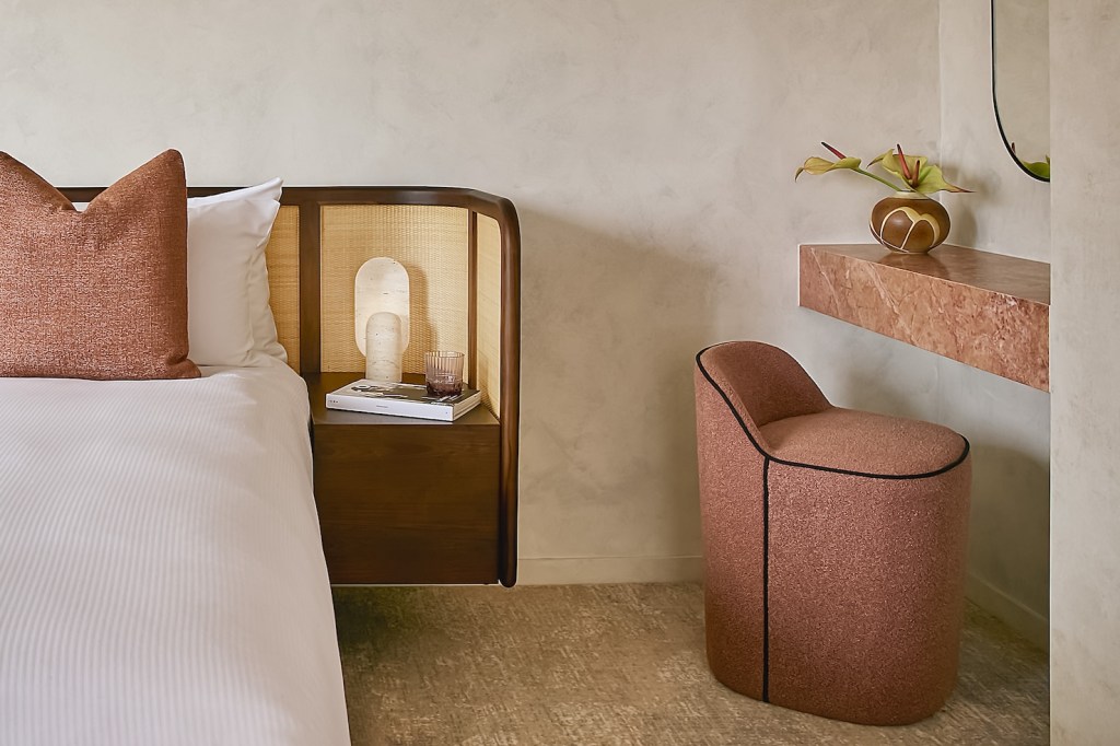 Corner of a white bed with pink pillow and pink vanity seat and vanity counter to the right