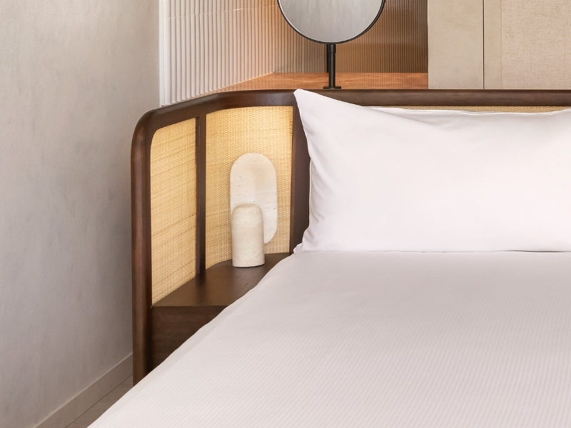 Close up image of white bed with wooden and weaved headboard and cream walls in the background