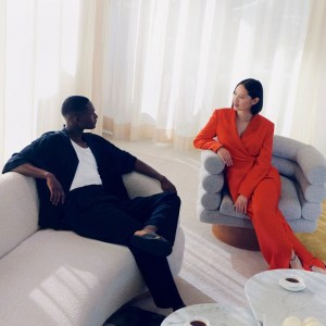 Woman in red suit sits on grey chair and man in black top and pants sits with legs crossed on grey sofa