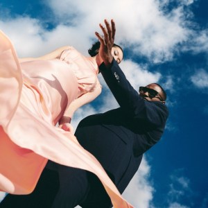 Man in dark suit and woman in peach dress pose in front of a cloudy blue sky