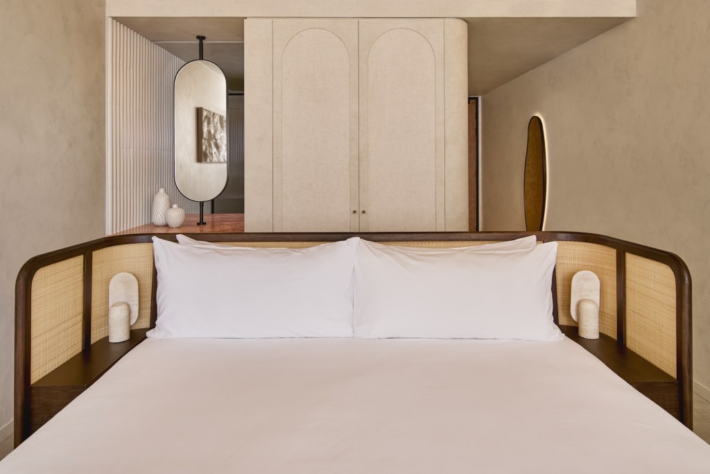 Close up image of a white bed with cream and dark wooden headboard and cream cabinet behind it and small mirror to the right