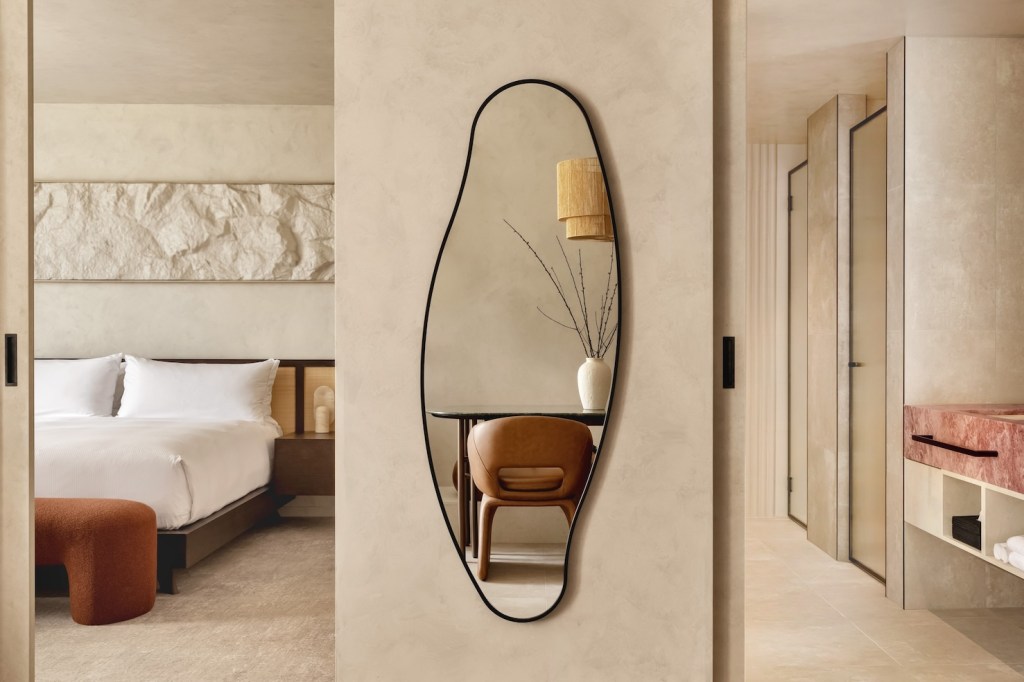 Suite with large white bed to the left, a mirror on the wall with twigs on a table in the reflection, and a cream and pink bathroom to the right