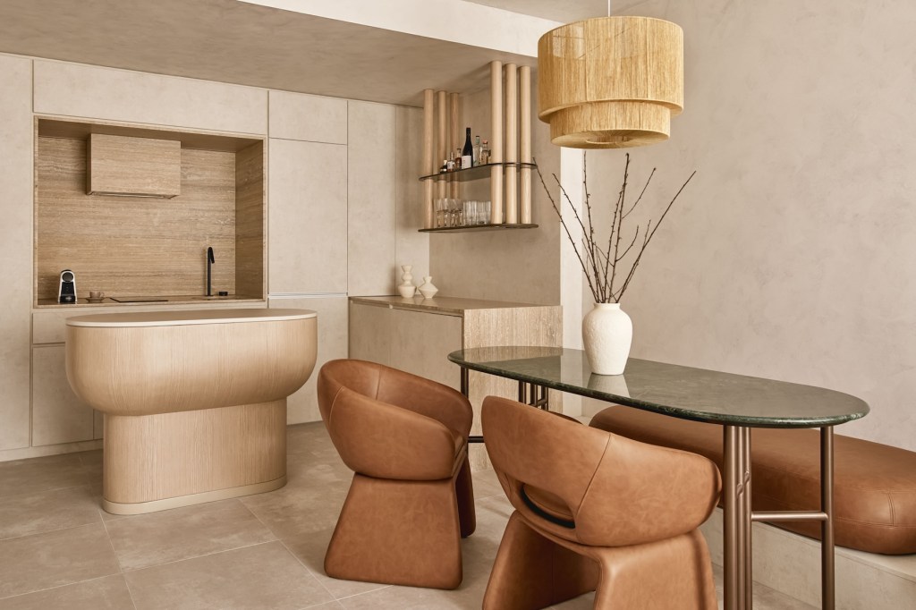 Dark oval table with pot and twigs on top and chestnut leather dining chairs surrounding it with light wooden kitchen island behind it