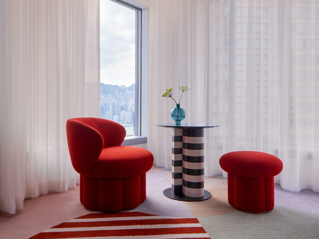 Red and white striped rug in a room with a red chair, red stool, small black and white table, and white curtains covering a window