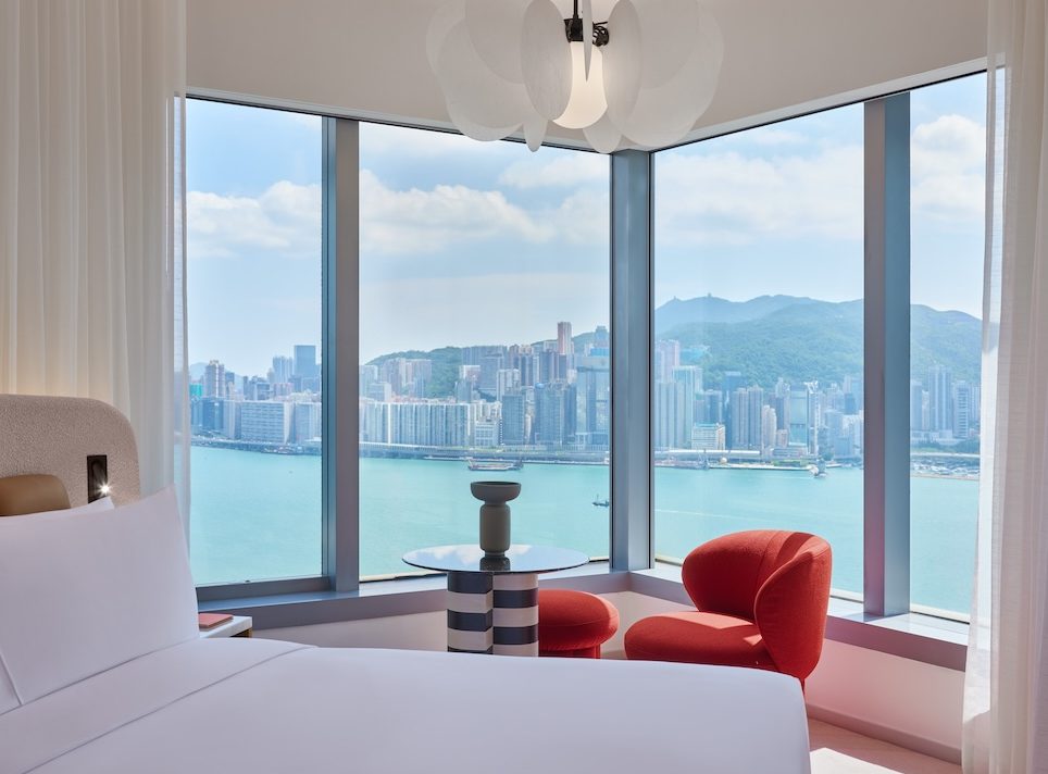 Side of white bed with window view of city and small table and red chair in the corner