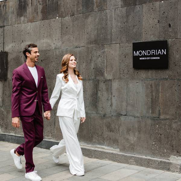 Man in purple suit and woman in white suit walk hand in hand in front of a grey building