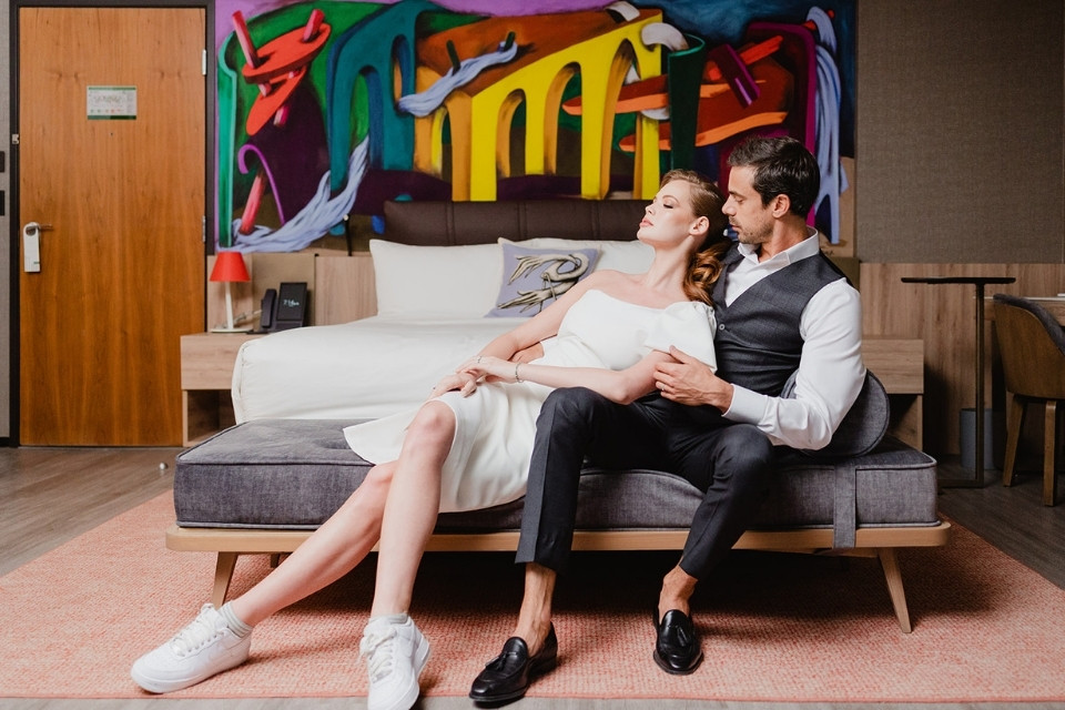 Man in suit and woman in white dress sit on the bench at the end of a bed embracing