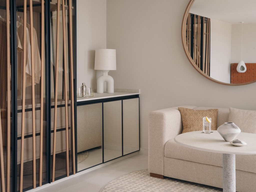 Mondrian Ibiza Studio Suite - Corner of a cream sofa with small pillow and white round coffee table with a round framed mirror on wall above and a glass hutch with lamp in the corner