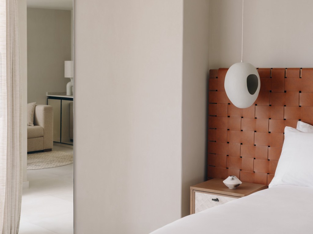 Mondrian Ibiza Studio Suite-Corner of a white bed with a rust-colored weaved headboard and a white hanging light fixture