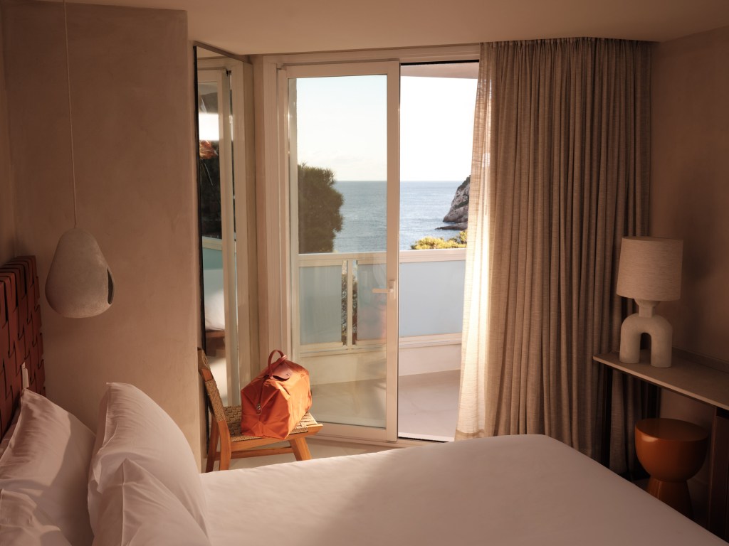 Good-Bedroom with natrual light seeping through the balcony windows, a large white bed in the middle, windows looking out to the sea, and an orange bag sitting on a bench