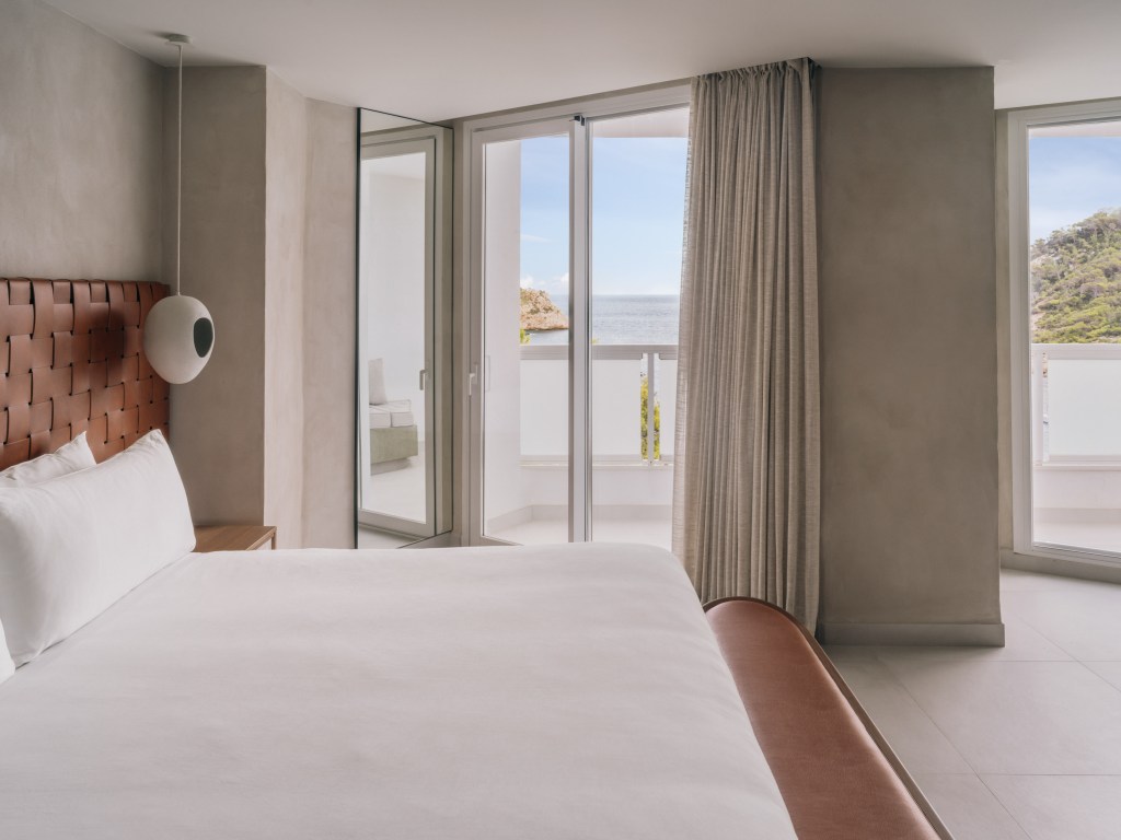 Mondrian Ibiza Panoramic Suite king size mattress which white sheets brown leather headboard with two bedside tables, a carpet and a modern lamps. Suite has a balcony looking towards sea views