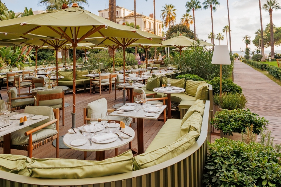 Outdoor restaurant with curved green banquette seats, green umbrellas, and lush palm trees and plants throughout