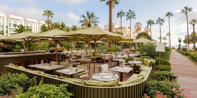 Beautiful garden restaurant with tables, chairs, and umbrellas set up with palm trees and a building in the distance