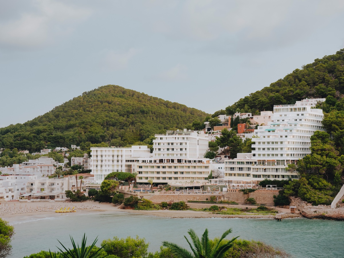 Large white building with green mountain behind it and sandy beach and turquoise water in front of it