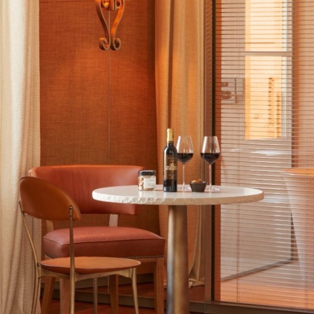 Two glasses of red wine and a bottle of red wine sit on a small round bistro table with two light brown chairs