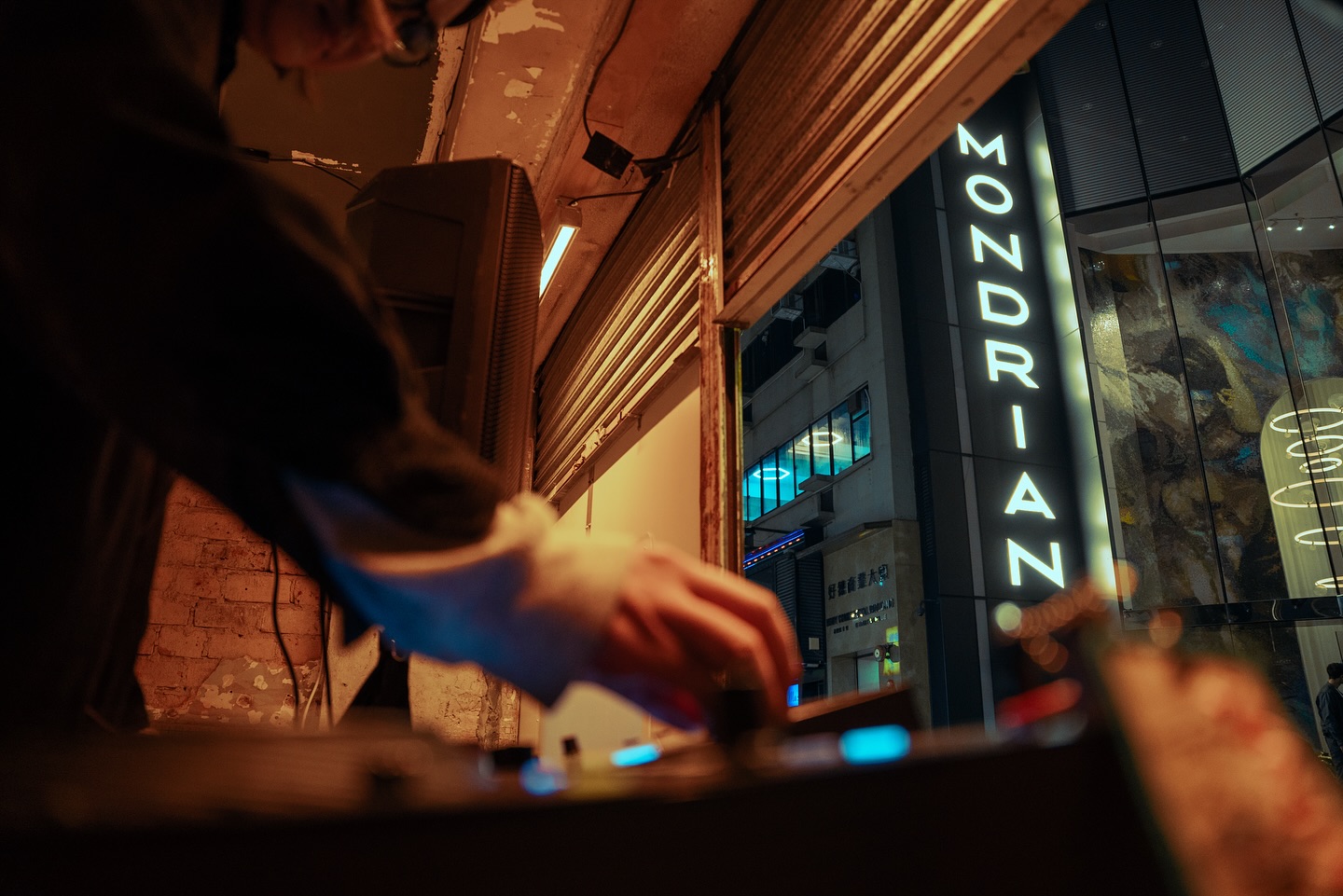 Man's hands touch a DJ keyboard in a garage like setting with lights of a Mondrian sign outside on a building