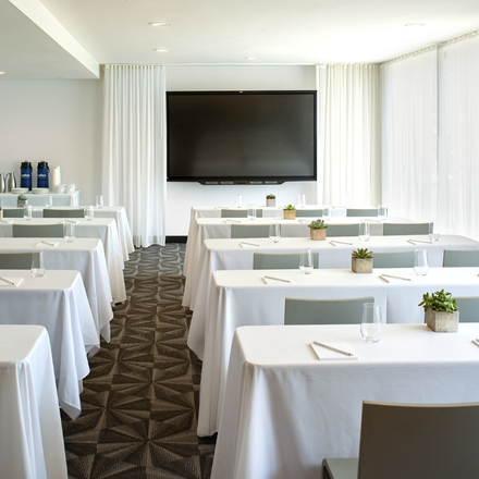 Rows of white covered tables and grey chairs and a tv screen at the front of the room