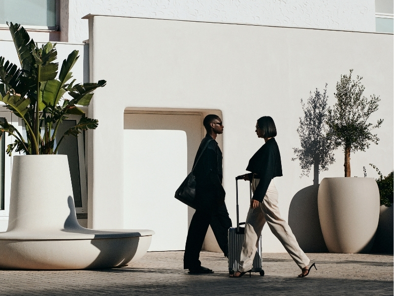 Woman with suitcase and man with bag walk outside a white building